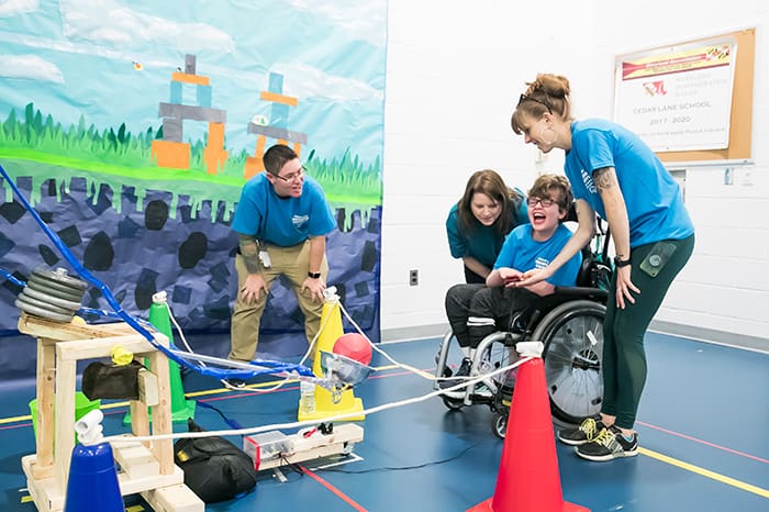 A male student sitting in a wheelchair and surrounded by three teachers is smiling as he plays with an adapted game.