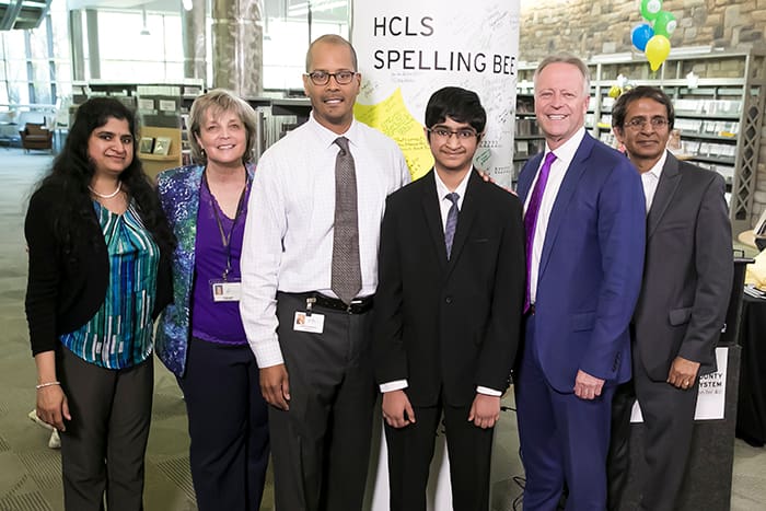 HCPSS Superintendent Matirano, Saketh Sundar, and a group of men and women stand in front of an HCLS Spelling Bee sign.