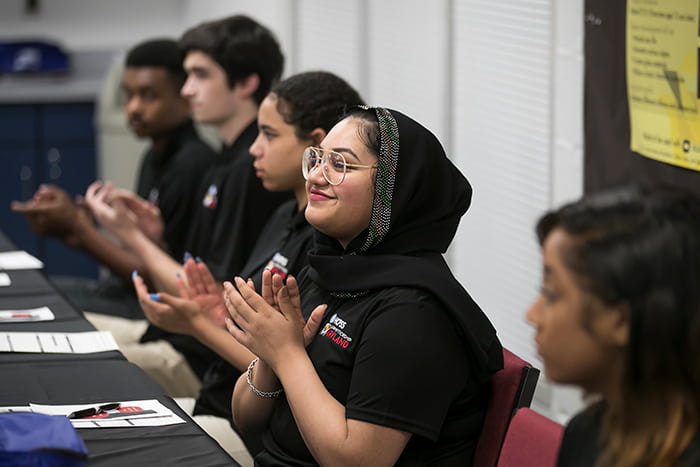 A female student seated at a table claps her hands alongside other seated students.