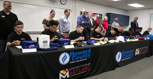 Students sit at a table and sign documents while a group of adults stands behind them.