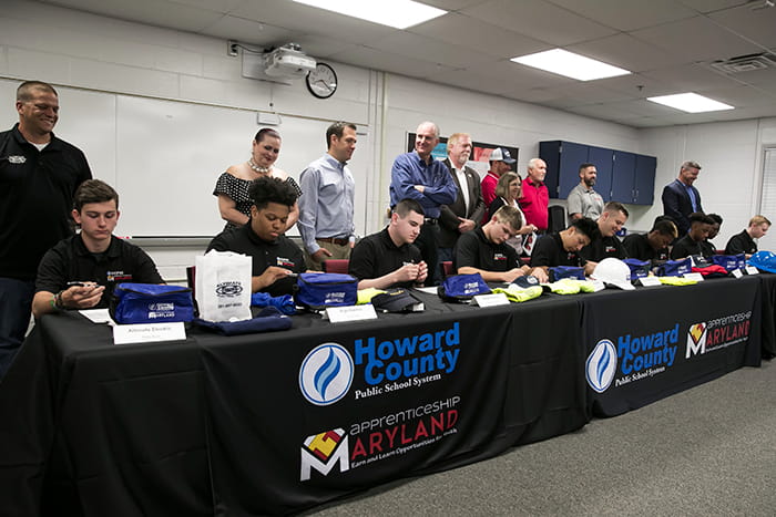 Students sit at a table and sign documents while a group of adults stands behind them.