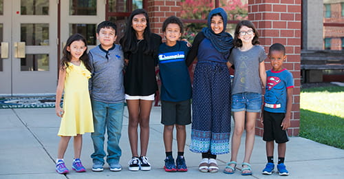 A diverse group of elementary school students locking arms together and standing in front of an HCPSS school.