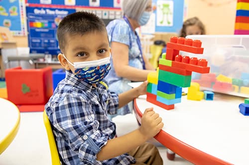 A younger, masked Prekindergarten student playing with Duplo blocks.
