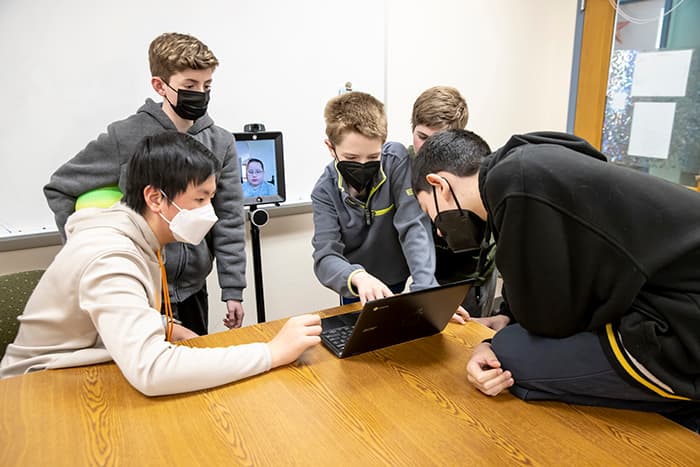 EMMS students, incuding a home and hospital student tuning in using a robot, gather around a computer screen.