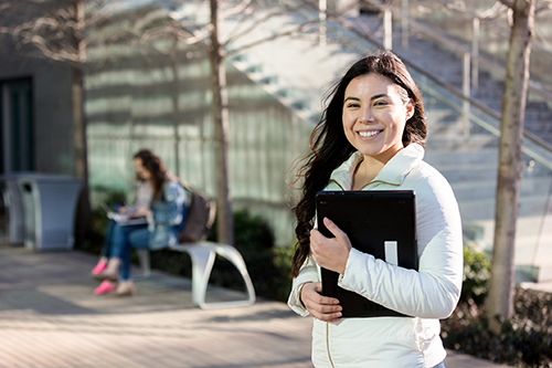 female standing outside, smiling and holding a laptop