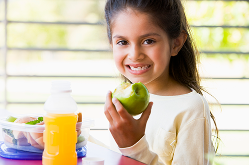 a young girl smiling and holding an apple. she is eating lunch with a sandwich and drink on the table.