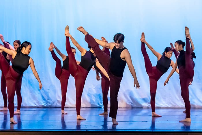Students in the All-County Dance Ensemble holding their legs in the air on stage.