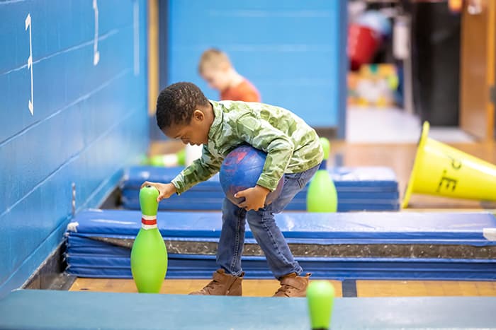 Male Laurel Woods ES pre-k student setting up bowling pins in the gym.