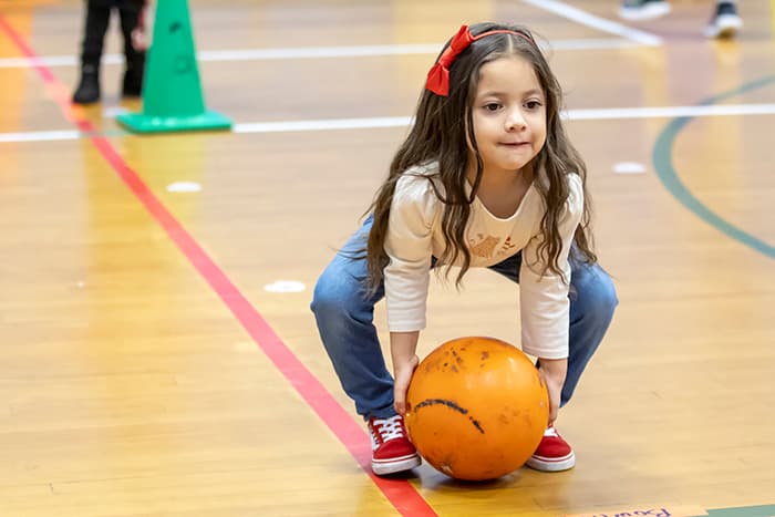 Female Laurel Woods ES pre-k student rolling a ball in the gym.