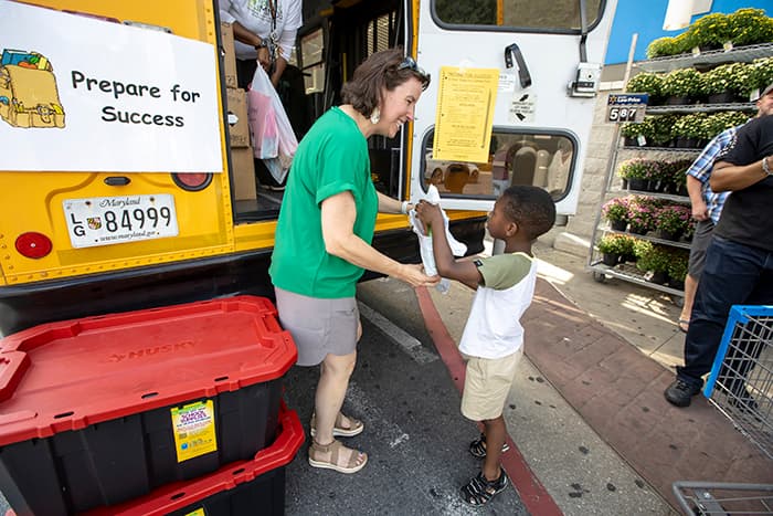 A young boy handing a bag of school supplies to a woman outside a school bus.