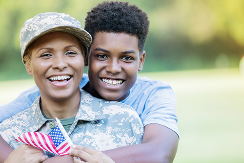 African American teenage boy stands behind his military mom, dressed in camo, and wraps his arms around her as they smile for the camera. She is holding a small American flag. 