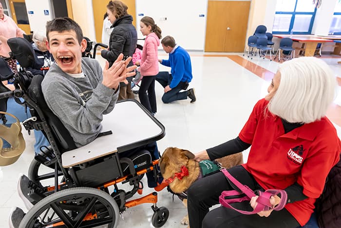 A student in a wheelchair claps and smiles while interacting with a dog.