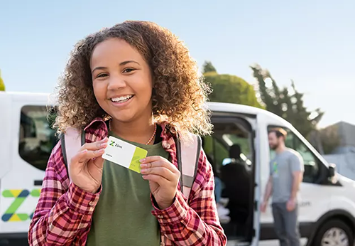 young girl smiling and holding a Zum RFID bus card. Blurred in background is Zum van with driver standing outside.