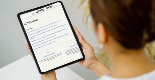 view from behind the shoulder of woman holding an ipad. on the ipad she is looking at the HCPSS News website