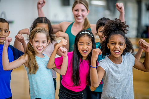 A group of elementary school students are posing with their teacher during gym class. They are all wearing athletic clothing, and flexing for the camera