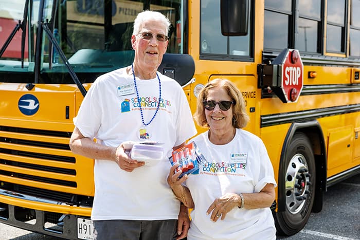 Roy and Susan Appletree holding school supplies in front of a school bus.