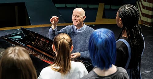 Greg Knauf sitting at a piano, conducting choral students.